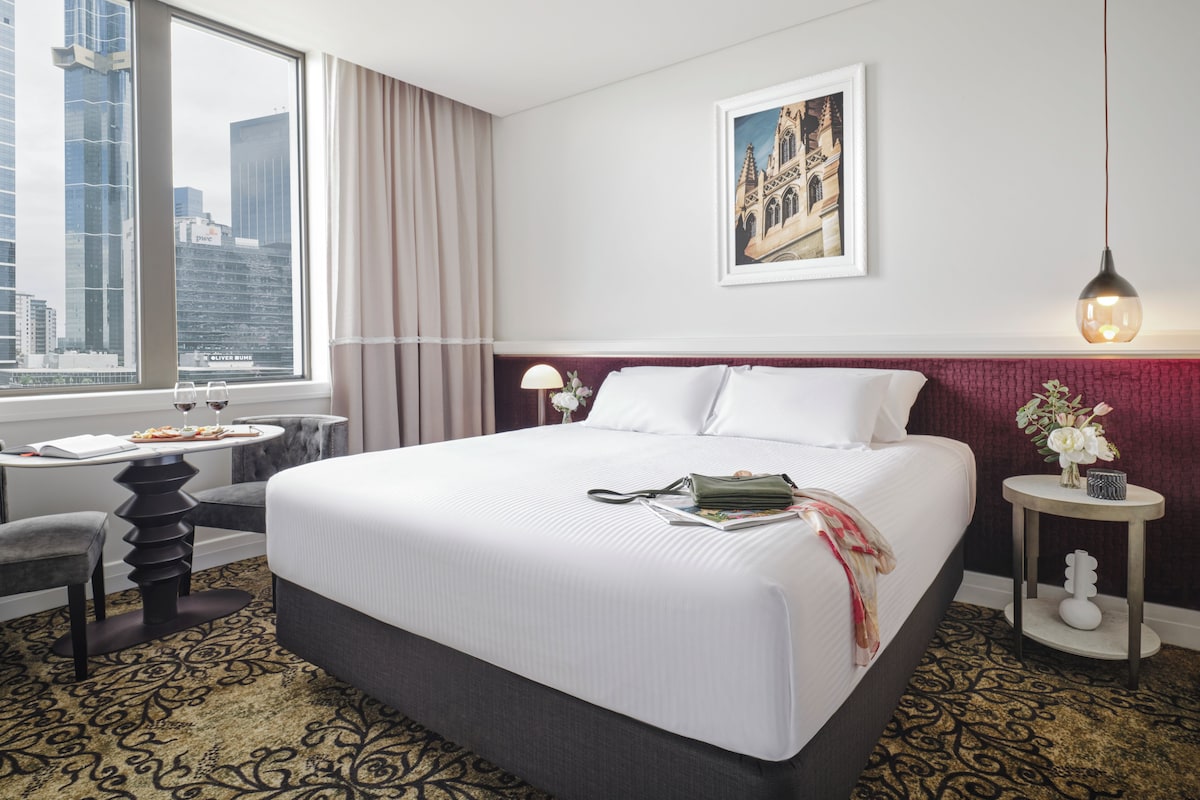 A hotel room with a large bed, a desk with a chair, a window with curtains, a framed picture on the wall, and a small table with a vase of flowers.