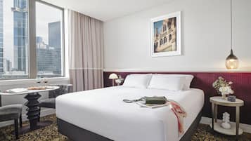 Habitación de hotel con una cama grande, un escritorio con silla, una ventana con cortinas, un cuadro enmarcado en la pared y una mesita con un jarrón de flores.