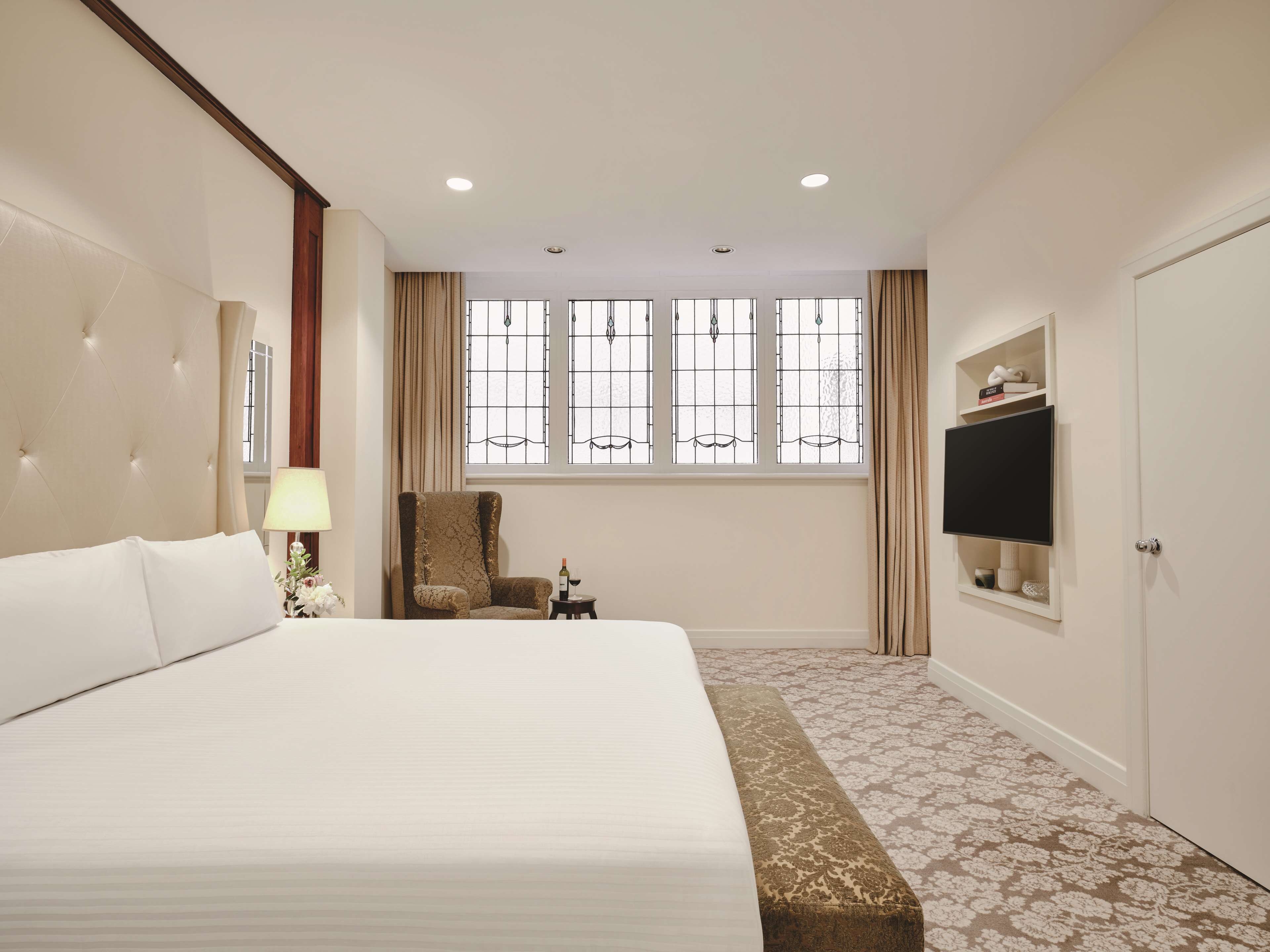 A hotel room with a large bed, a chair, a TV mounted on the wall, and a window with curtains.