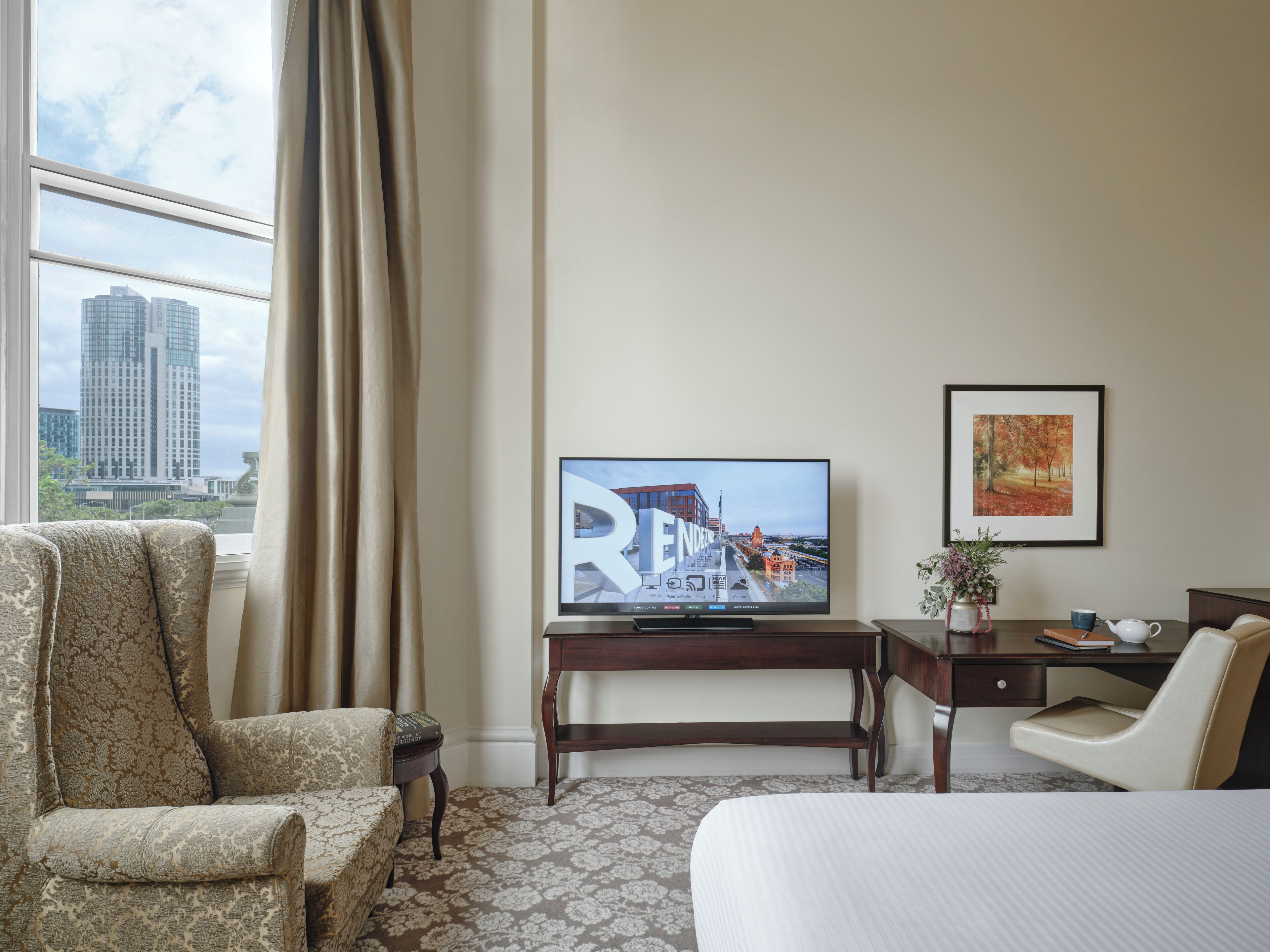 A hotel room with a bed, a chair, a desk with a television, a framed picture, and a view of a cityscape through a window.