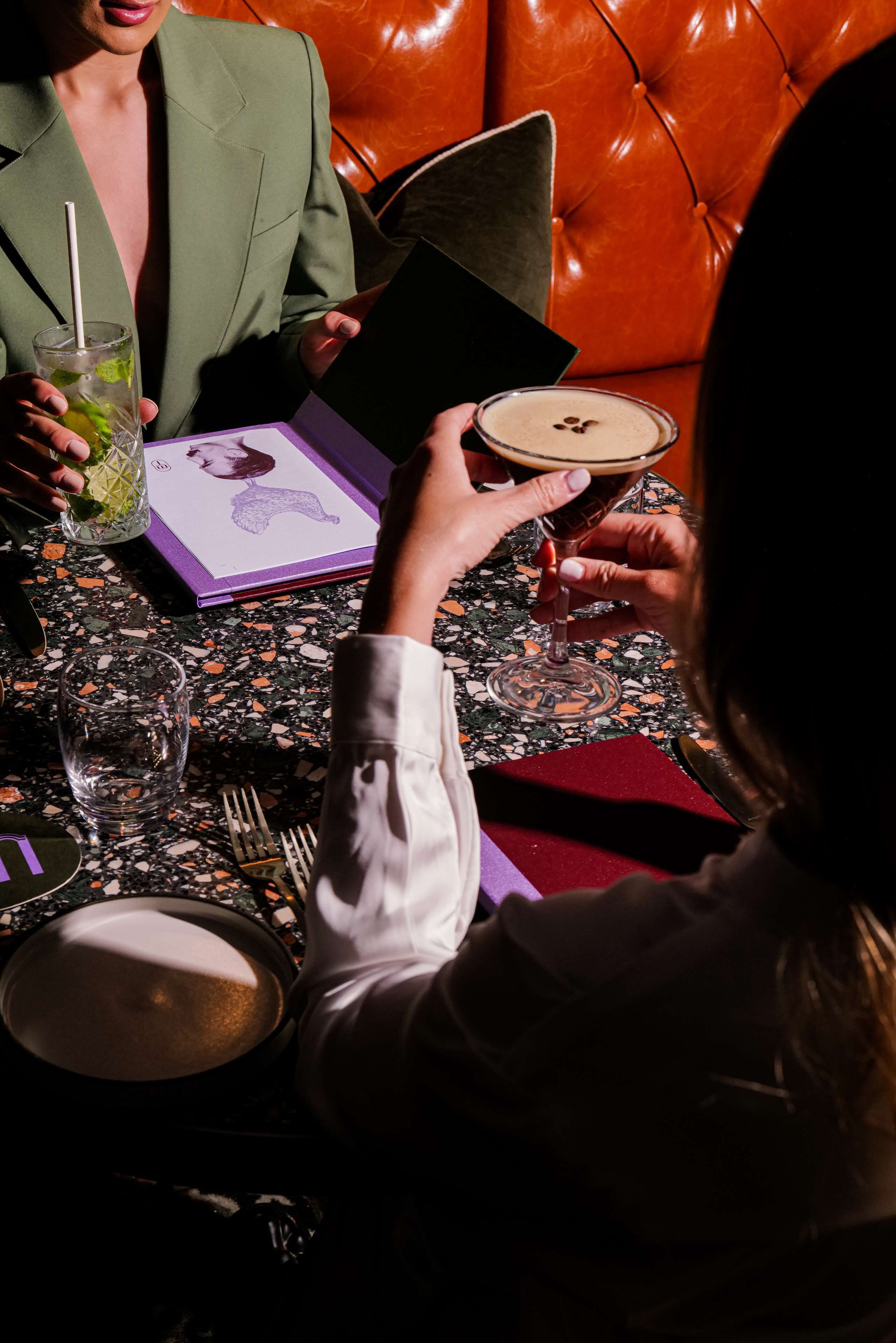 A person holding a menu at a restaurant with a leather booth, a glass of water, and a drink with a straw on the table.