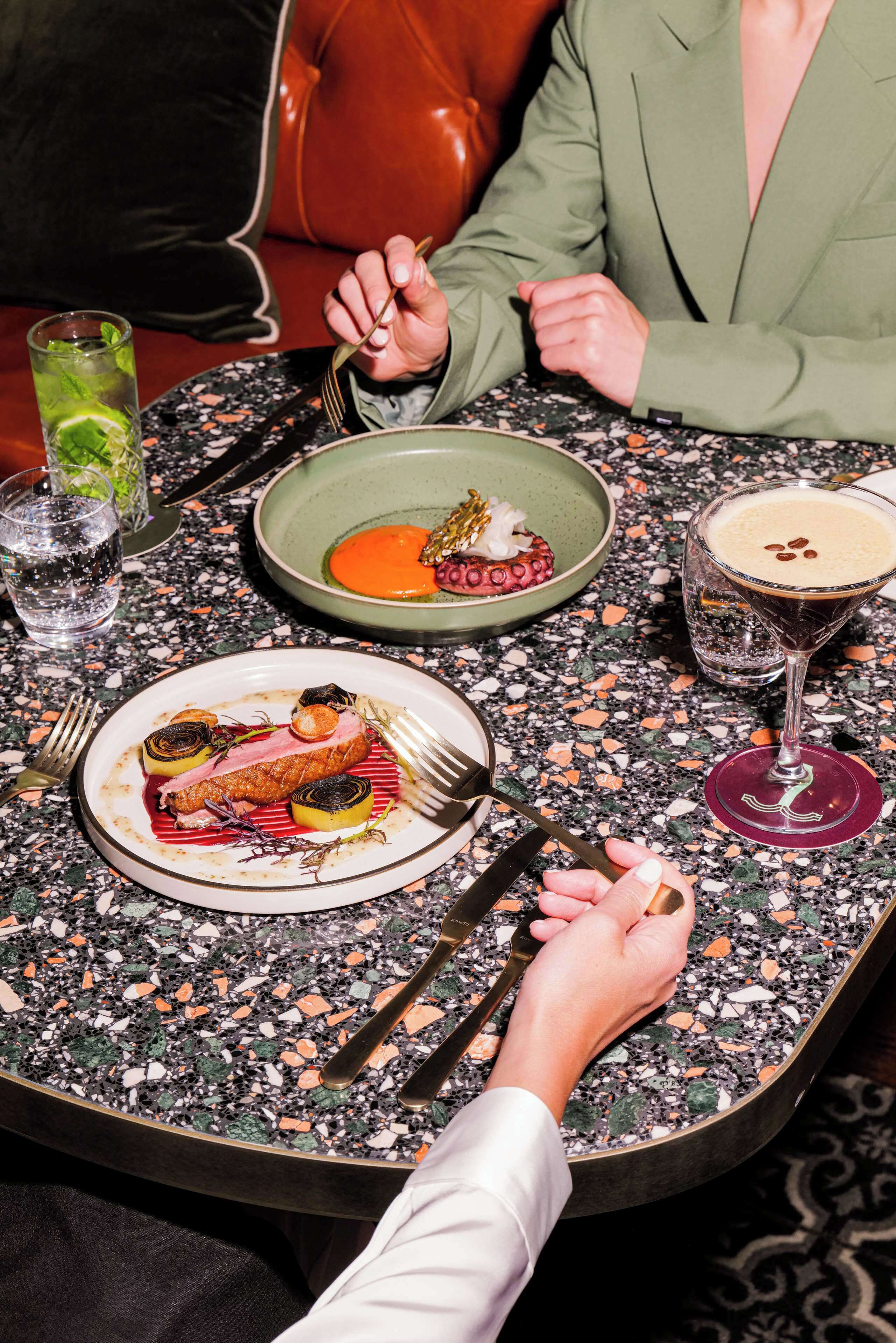 A dining table with a variety of dishes, including a plate with a piece of meat and vegetables, a bowl with a red sauce, and a glass of a dark-colored beverage.