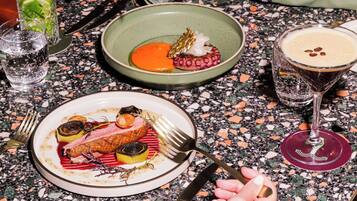 A dining table with a variety of dishes, including a plate with a piece of meat and vegetables, a bowl with a red sauce, and a glass of a dark-colored beverage.