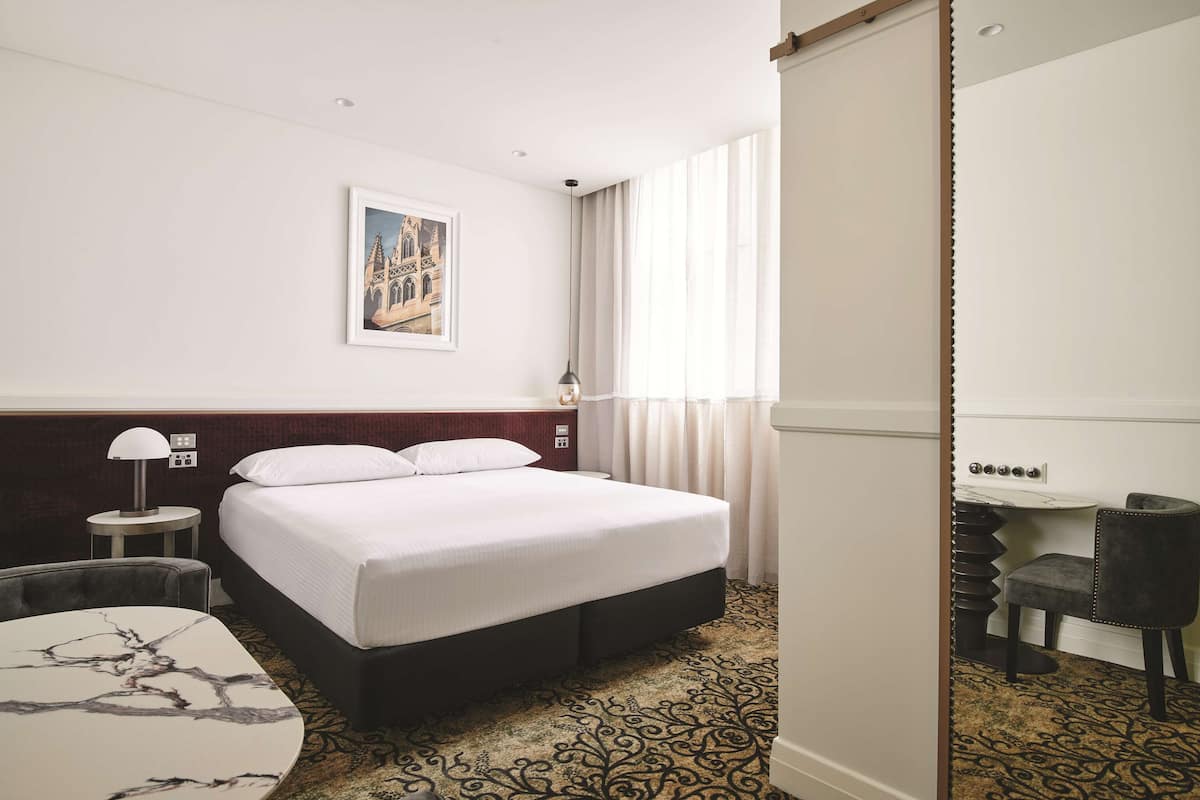 A hotel room with a large bed, a desk with a chair, a small table, and a patterned carpet.