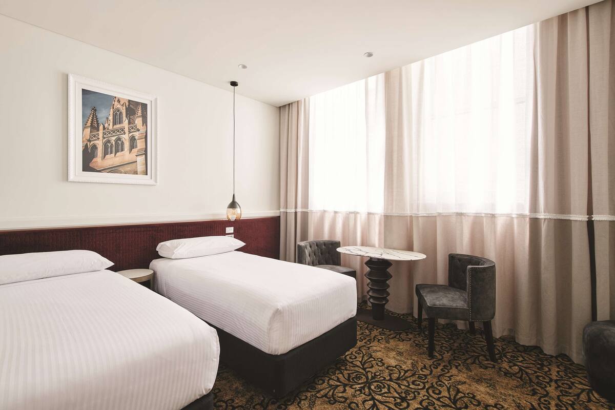 A hotel room with two beds, a small table, a chair, and a large window with curtains.
