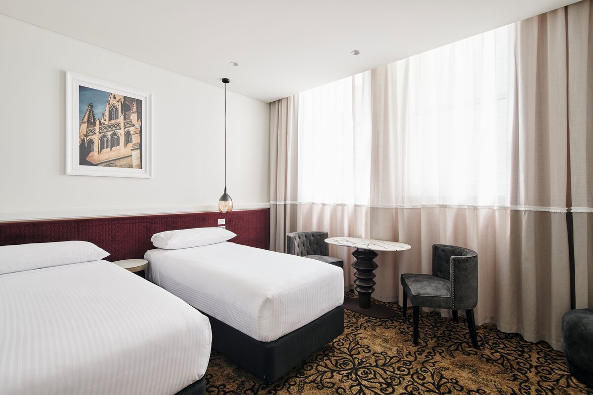 Habitación de hotel con dos camas, una mesita redonda pequeña, una silla y un amplio ventanal con cortinas.