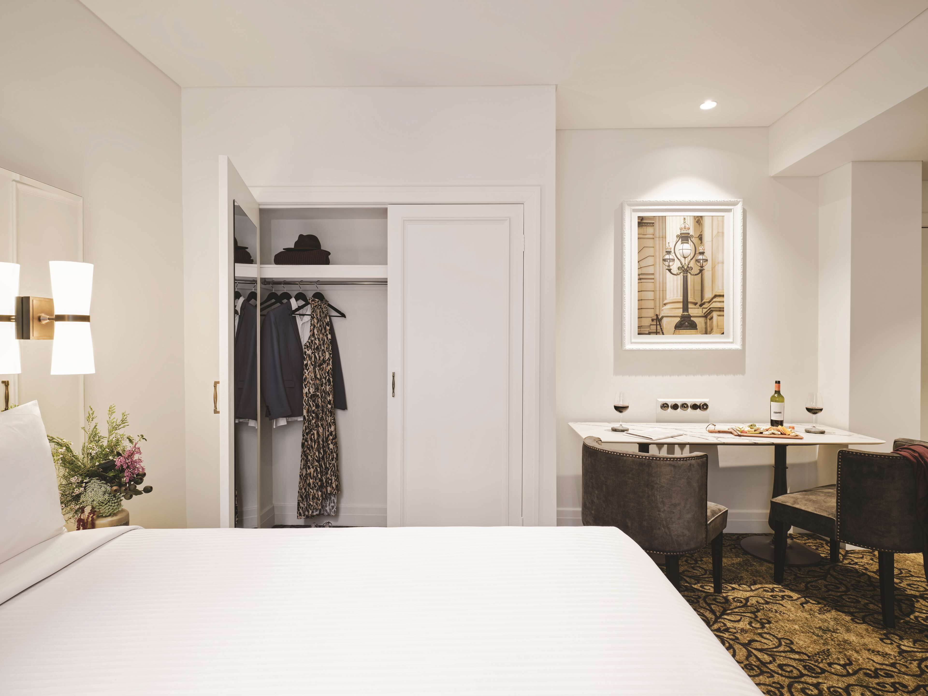 A hotel room with a bed, a desk with a chair, a closet with clothes, a framed picture on the wall, and a vase with flowers on the nightstand.