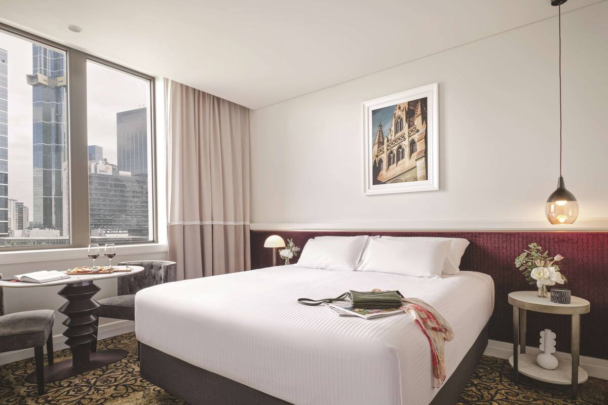 A hotel room with a large bed, a desk with a chair, a window with curtains, a framed picture on the wall, and a hanging light fixture.
