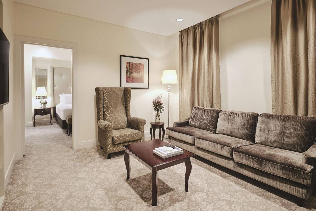 A hotel room with a large bed, a sofa, a coffee table, a lamp, a framed picture on the wall, and a television.