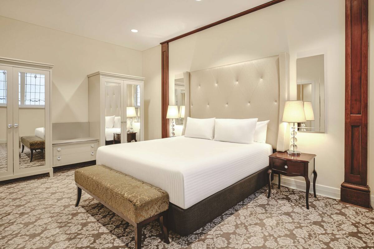 A spacious bedroom with a large bed, a wardrobe, a bench, and a nightstand with a lamp.