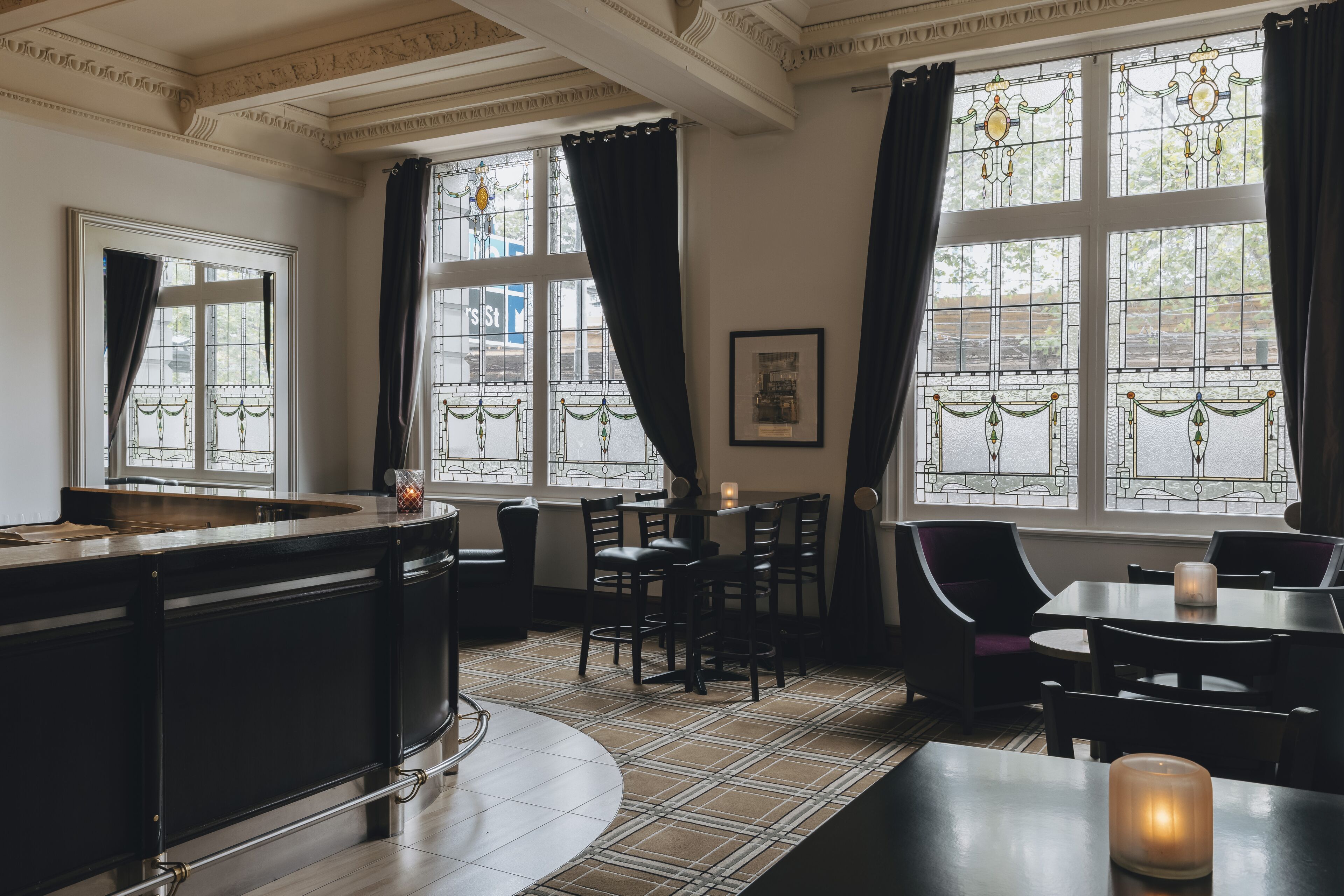 A restaurant with a bar area, dining tables, chairs, and large windows with curtains.