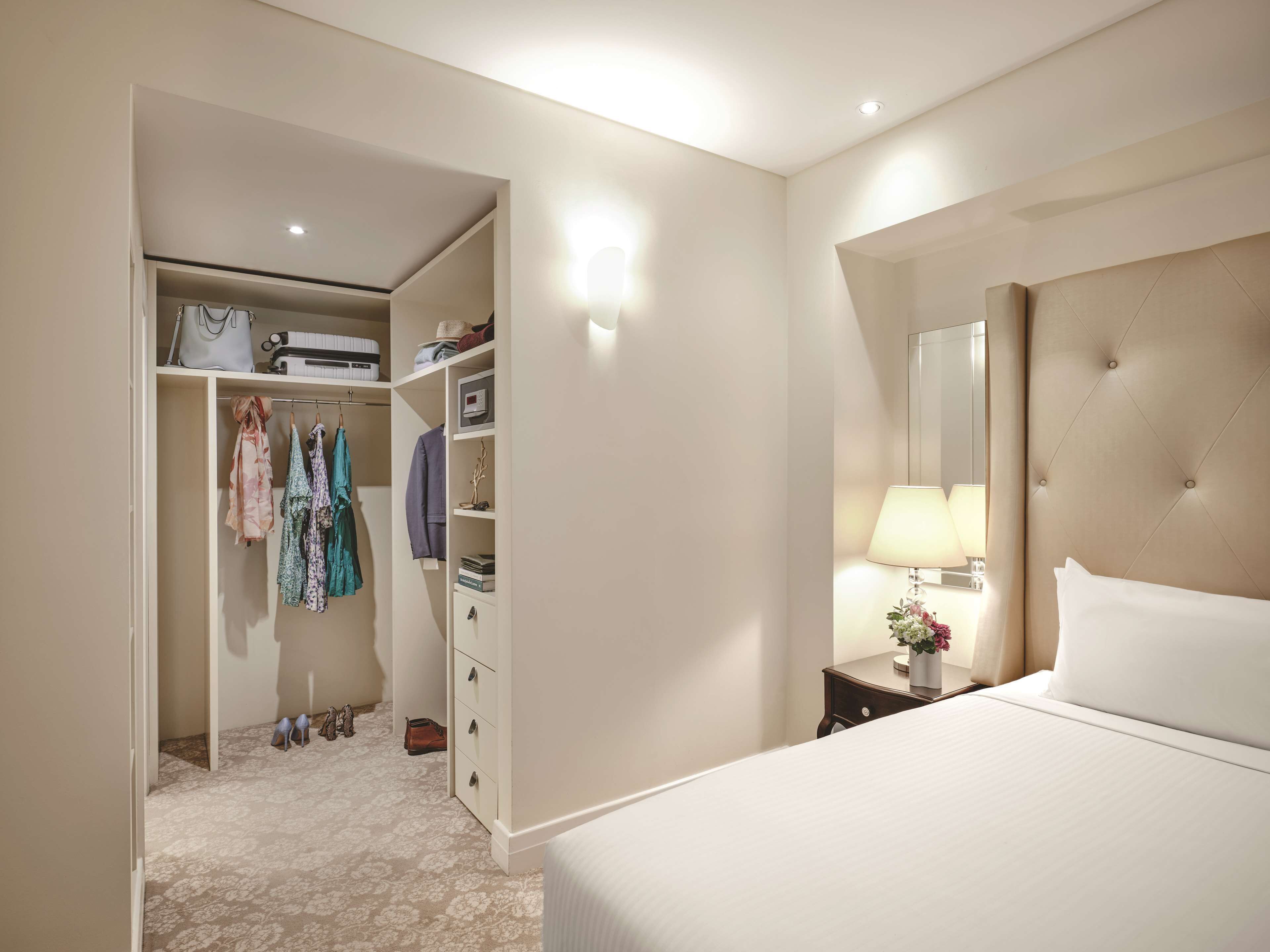 A hotel room with a bed, a bedside table with a lamp, a large mirror, a wardrobe, and a shoe rack.