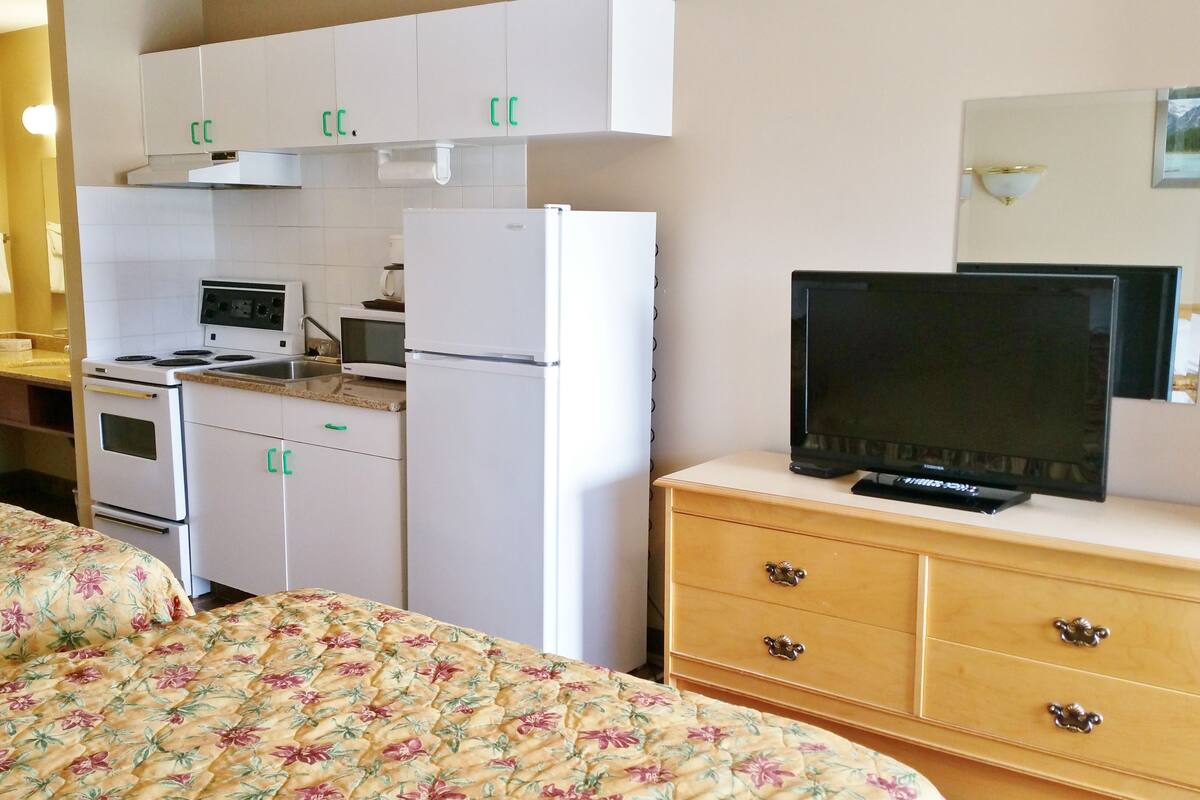 Une chambre d’hôtel avec une kitchenette, un lit avec une literie à motifs floraux, une télévision et une commode en bois.
