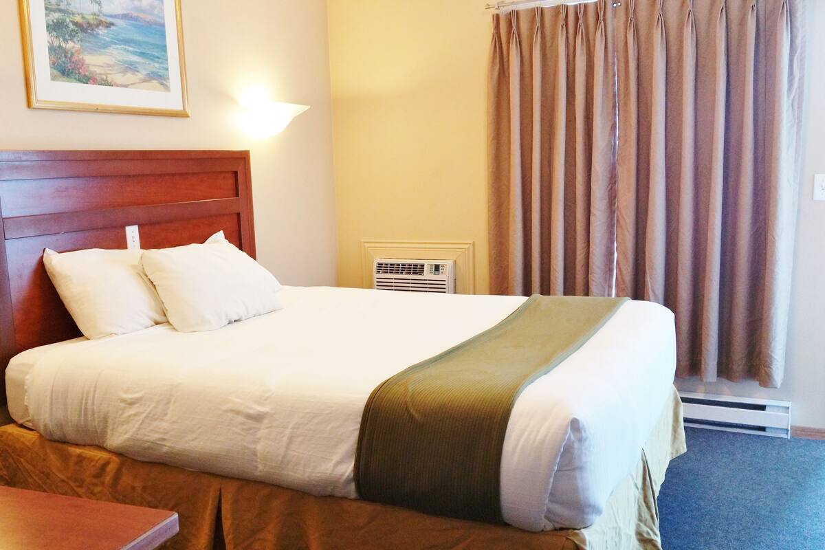 Une chambre d’hôtel avec un lit, une table de chevet, une lampe, un tableau et une fenêtre avec des rideaux.