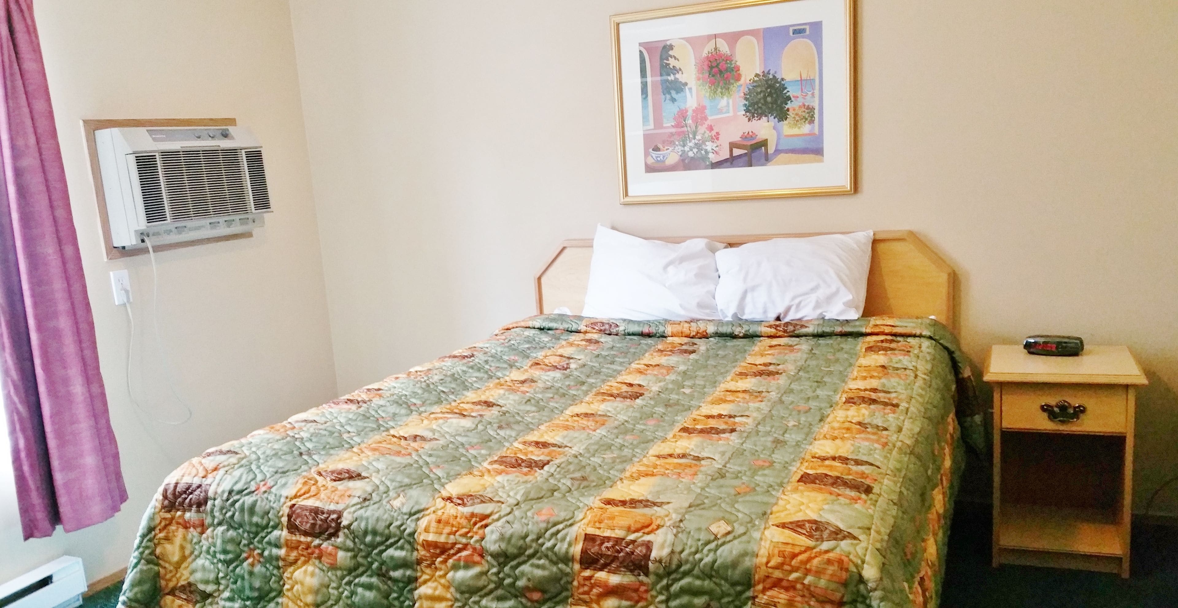 Uma cama bem arrumada com um edredom, uma mesinha de cabeceira com uma garrafa, e um ar condicionado fixado na parede.