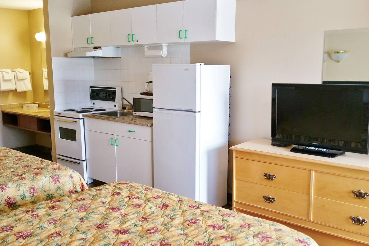 Une chambre d’hôtel comprenant un lit, une télévision, une kitchenette équipée d’une cuisinière, d’un four à micro-ondes et d’un réfrigérateur, ainsi qu’une commode en bois.