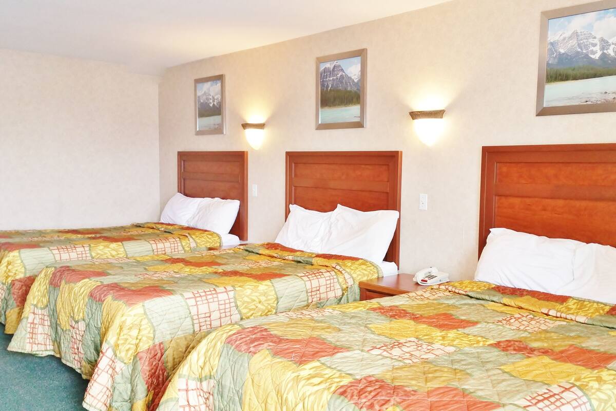Une chambre d’hôtel avec trois lits, chacun doté d’une tête de lit en bois et d’une couverture colorée. Des lampes murales et des tableaux encadrés sont accrochés au mur au-dessus des lits.
