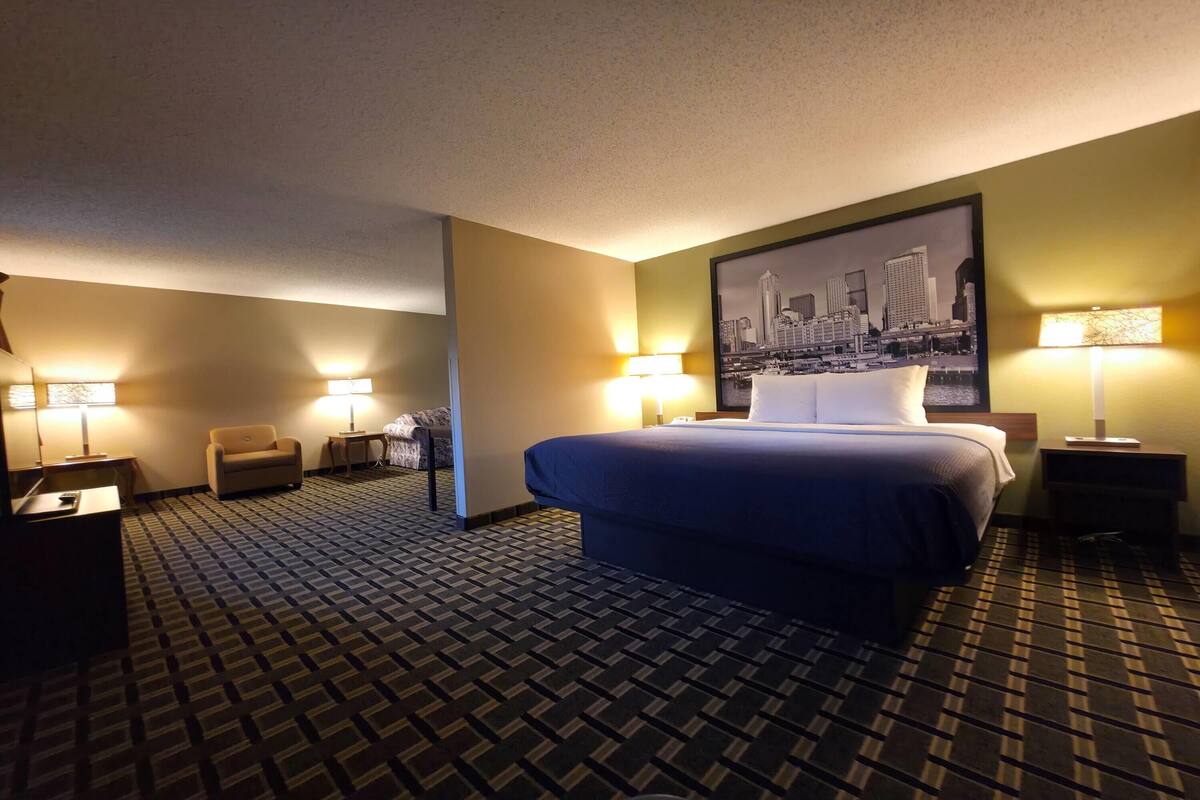 A hotel room with a bed, two bedside tables, a chair, a desk, and a cityscape painting on the wall.