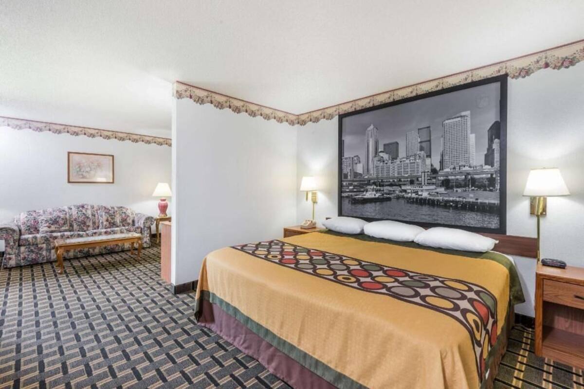 A hotel room with a bed, a sofa, a cityscape painting, and a patterned carpet.
