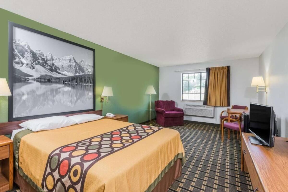 A hotel room with a bed, a desk with a computer, a chair, a TV, a window with curtains, and a painting on the wall.