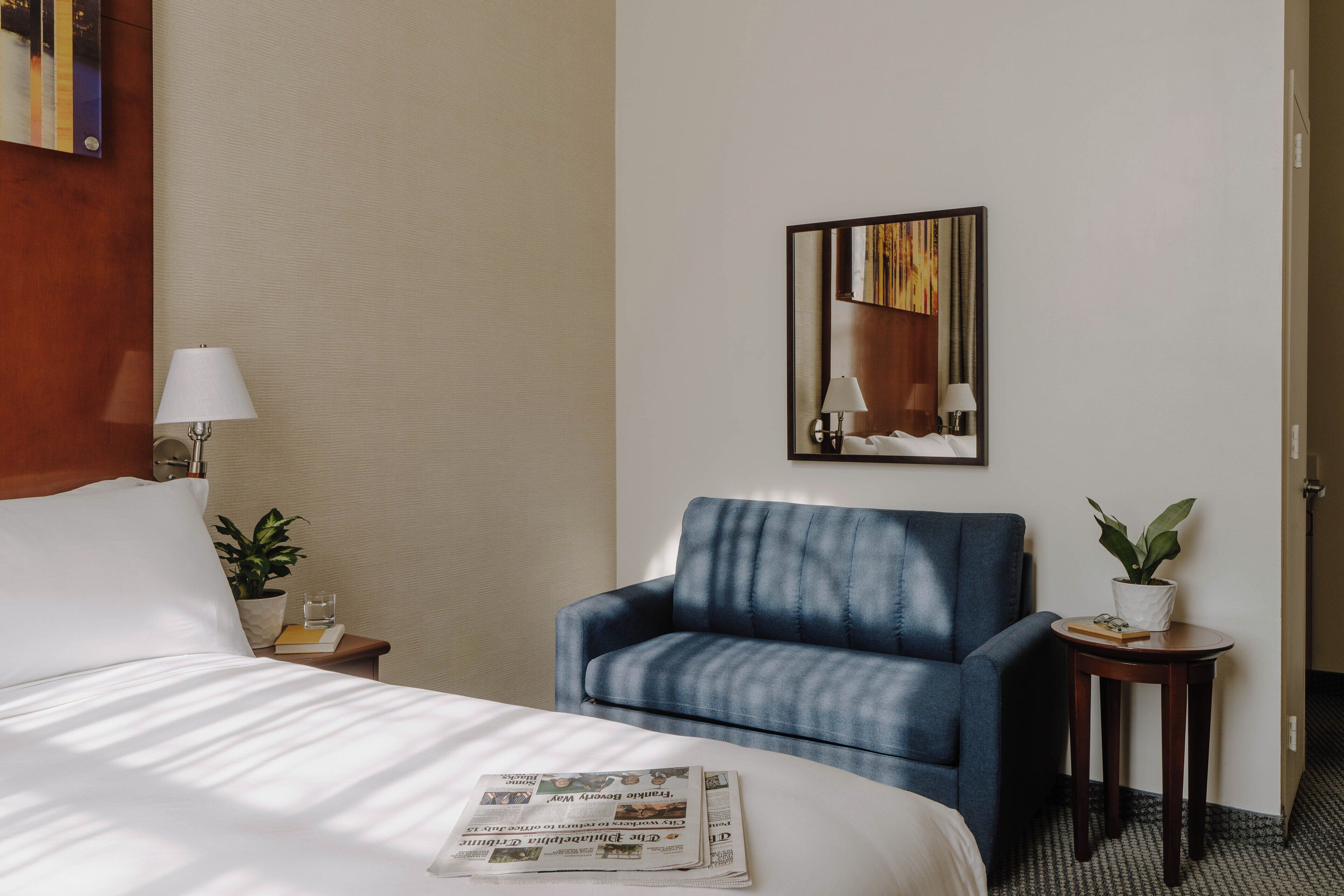 Quarto de hotel com uma cama, uma poltrona azul, uma pequena mesa com uma planta e um espelho fixado na parede.