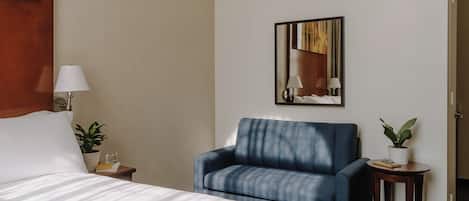 Habitación de hotel con una cama, un sillón azul, una mesita con una planta y un espejo en la pared.