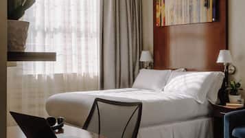 Una habitación de hotel con una cama, un escritorio con una computadora portátil, una silla, una planta en maceta y un cuadro en la pared.