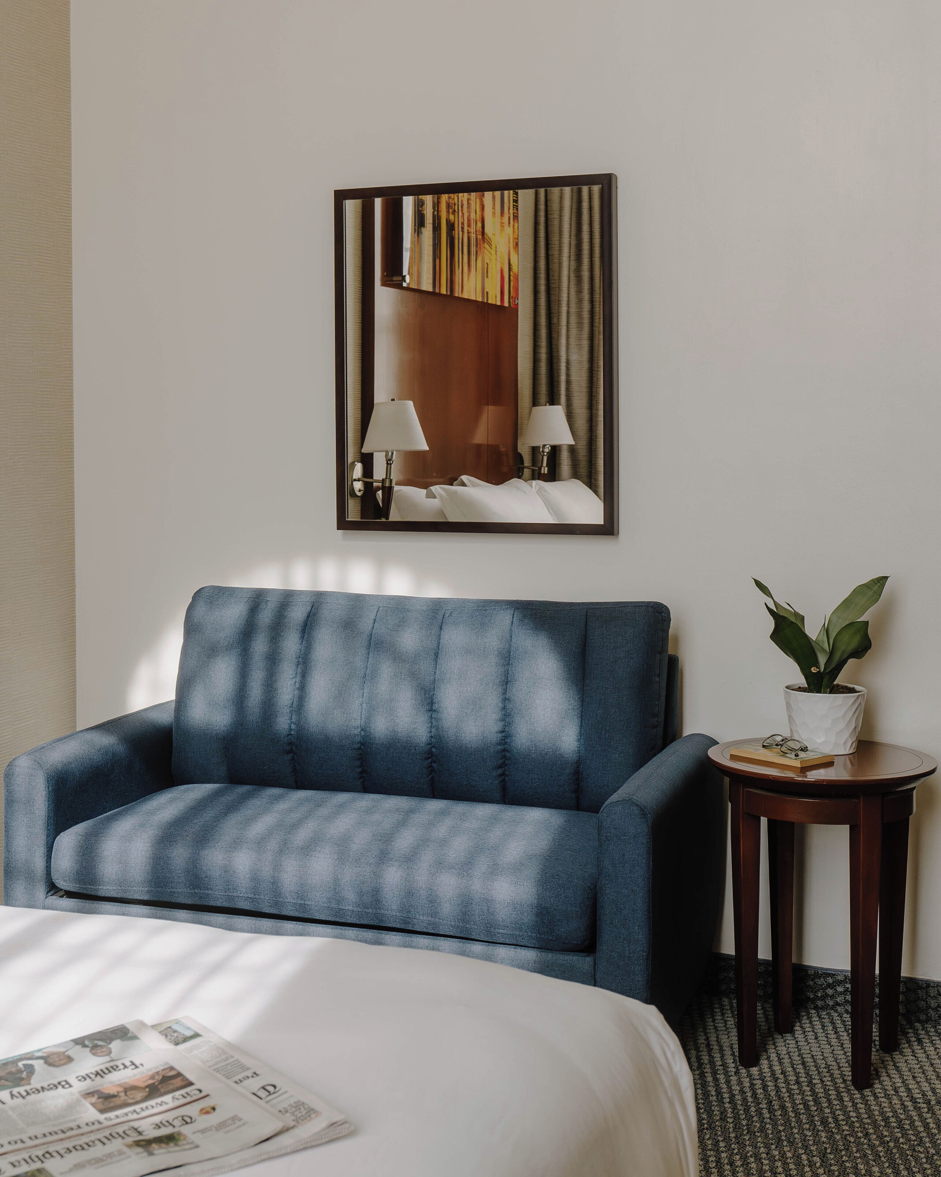 A hotel room with a bed, a blue sofa, a small table with a plant, and a mirror on the wall.