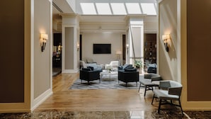 A spacious living area with a high ceiling and skylight, featuring a central coffee table surrounded by sofas and armchairs, with a bookshelf and a flat-screen TV in the background. - Club Quarters Hotel Rittenhouse Square, Philadelphia (Philadelphia)