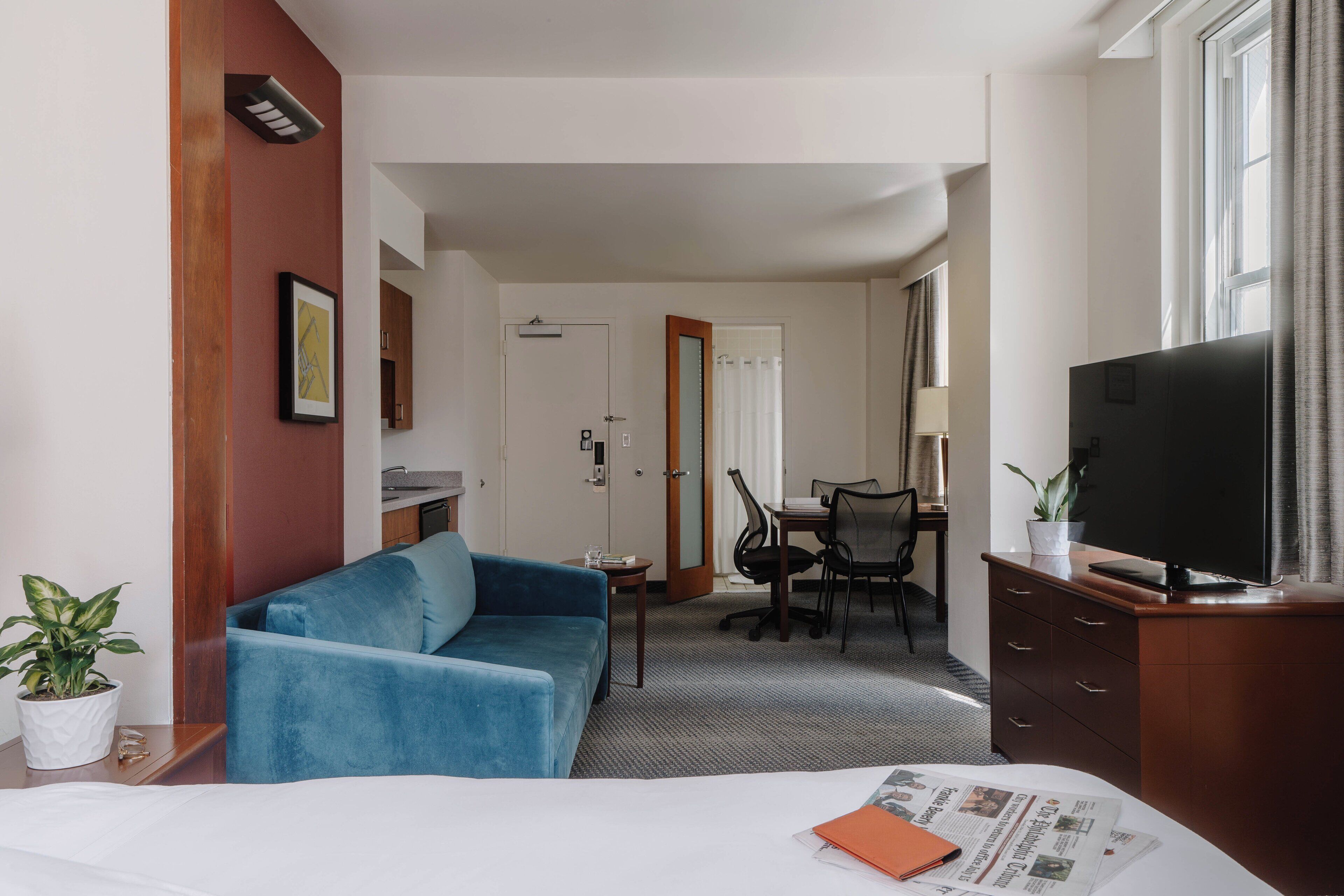 A hotel room with a bed, a blue sofa, a TV, a dining table with chairs, and a small table with a plant.