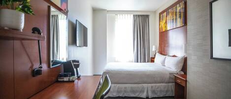 A hotel room with a bed, a desk, a chair, a TV, a plant, and a painting on the wall.