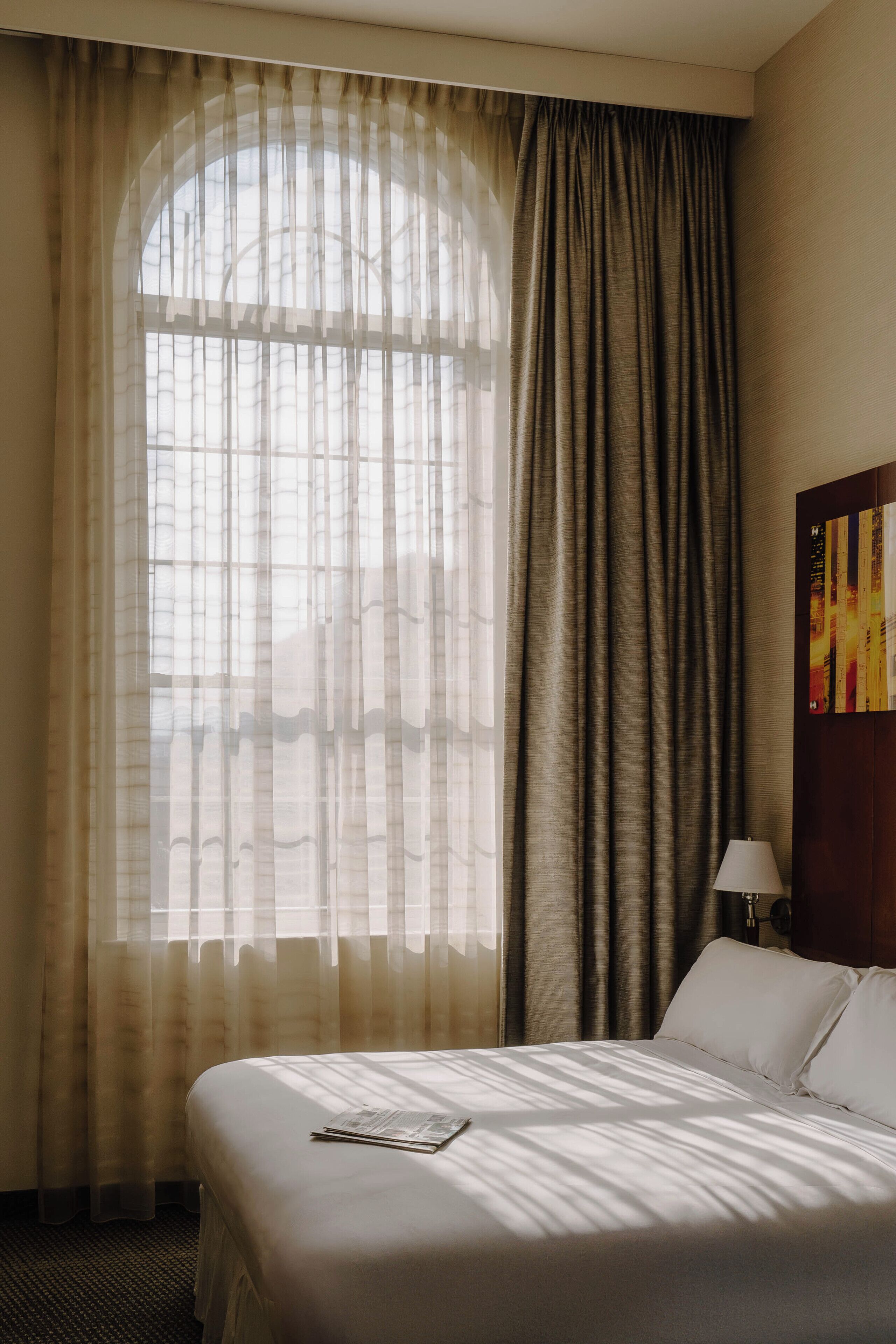Een hotelkamer met een groot bed, een raam met dunne gordijnen en een schilderij aan de muur.