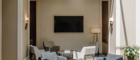 A spacious living area with a flat-screen TV mounted on the wall, surrounded by modern furniture and a large window providing natural light.