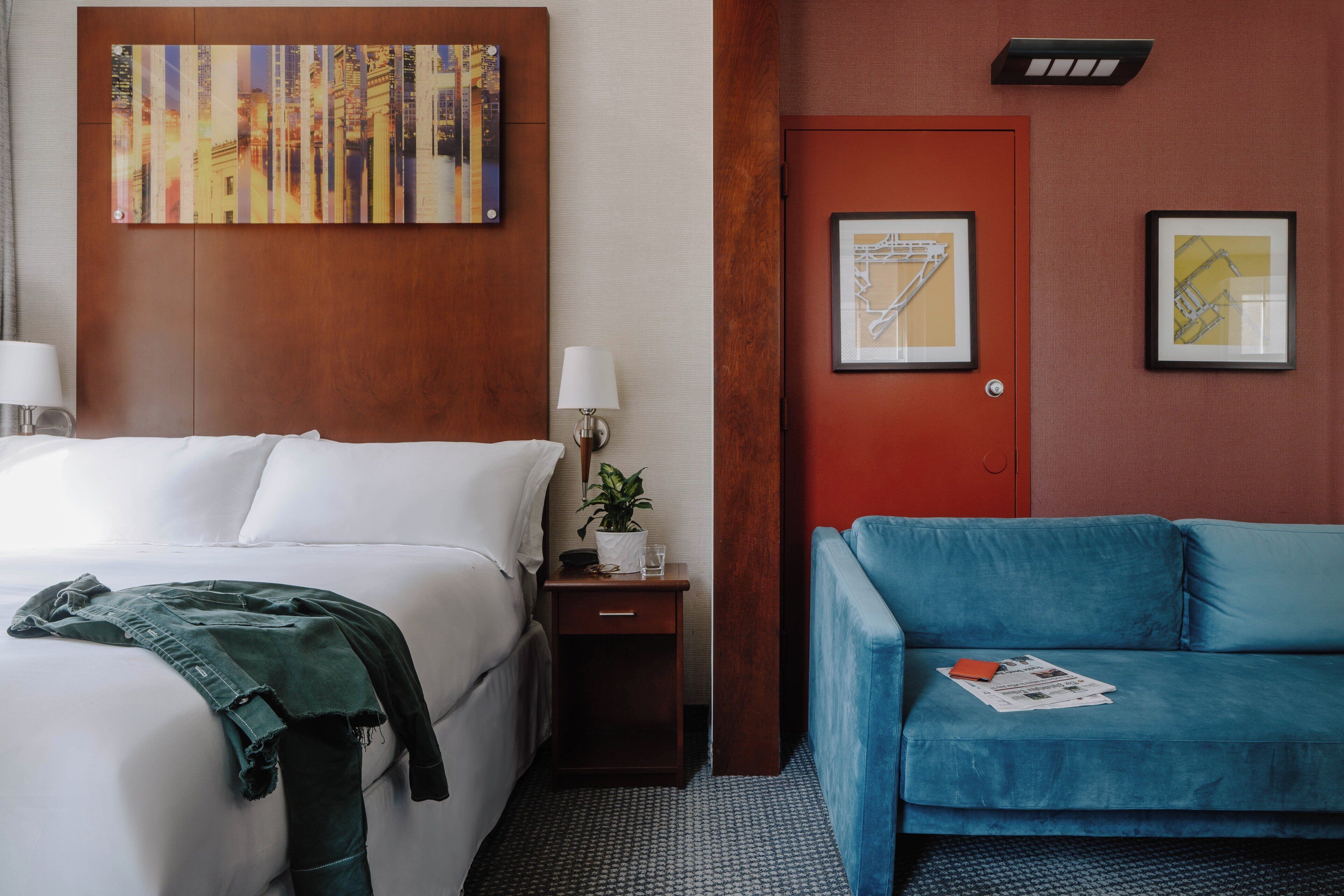 A hotel room with a bed, a blue sofa, a nightstand with a lamp, a door, and two framed pictures on the wall.