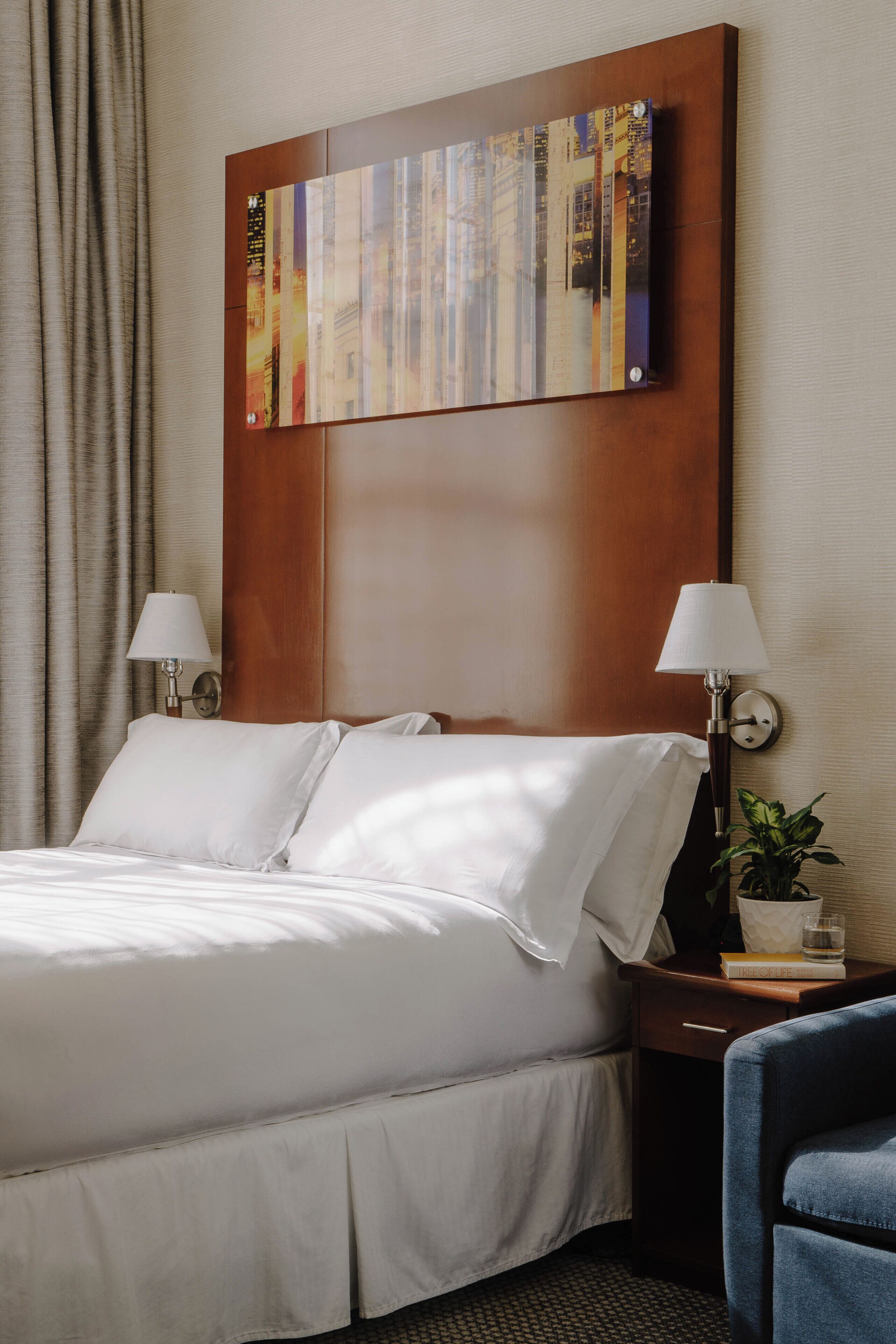 A hotel room with a large bed, two bedside lamps, a nightstand with a plant, and a wall-mounted artwork.