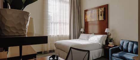 Camera d'albergo con un letto, una scrivania con un portatile, una sedia, un divano blu, un comodino con una lampada, una pianta in vaso e un quadro appeso al muro.
