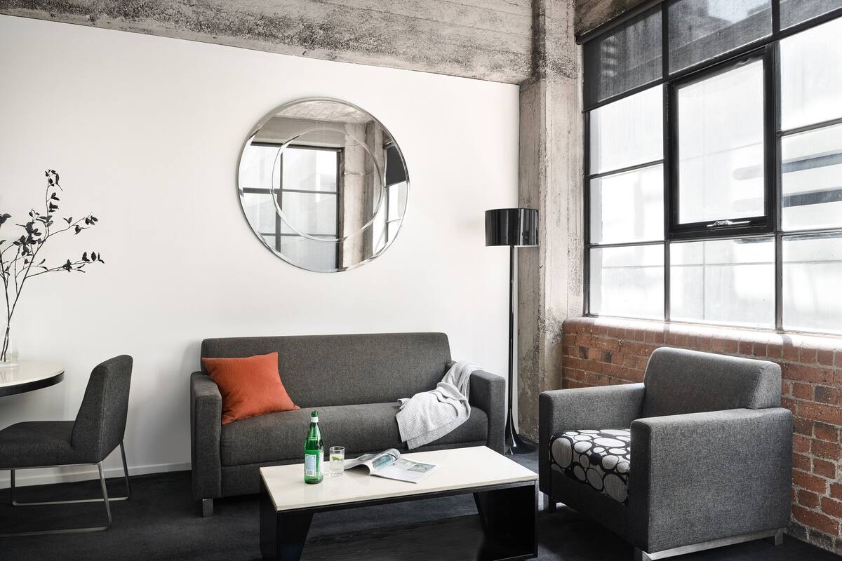 Ein modernes Zimmer mit einem grauen Sofa, einem runden Spiegel, einem Tisch mit Flasche und Zeitschrift sowie einem Fenster mit Aussicht.