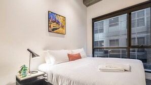 A hotel room with a large bed, a nightstand with a lamp, a painting on the wall, and a window with a view of buildings. - Punthill Manhattan (Melbourne)
