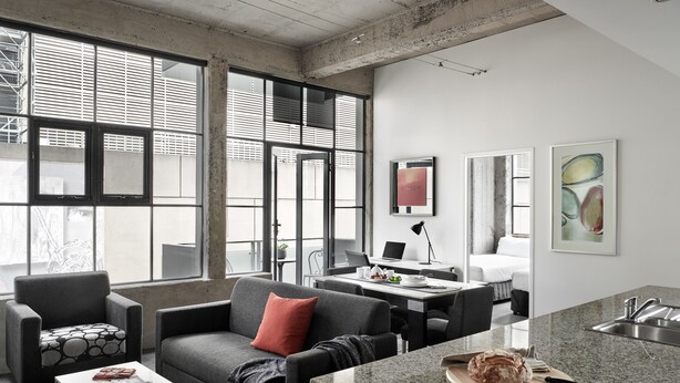 A modern living room with a sofa, armchair, and coffee table, a kitchen area with a granite countertop and a loaf of bread, and large windows with blinds. - Punthill Manhattan (Melbourne)