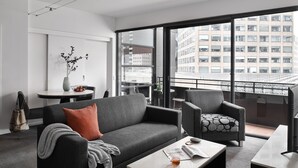 A modern living room with a grey sofa, a patterned armchair, and a coffee table. There is a vase with branches on the wall, a dining table with chairs, and a balcony with a view of buildings. - Punthill Manhattan (Melbourne)