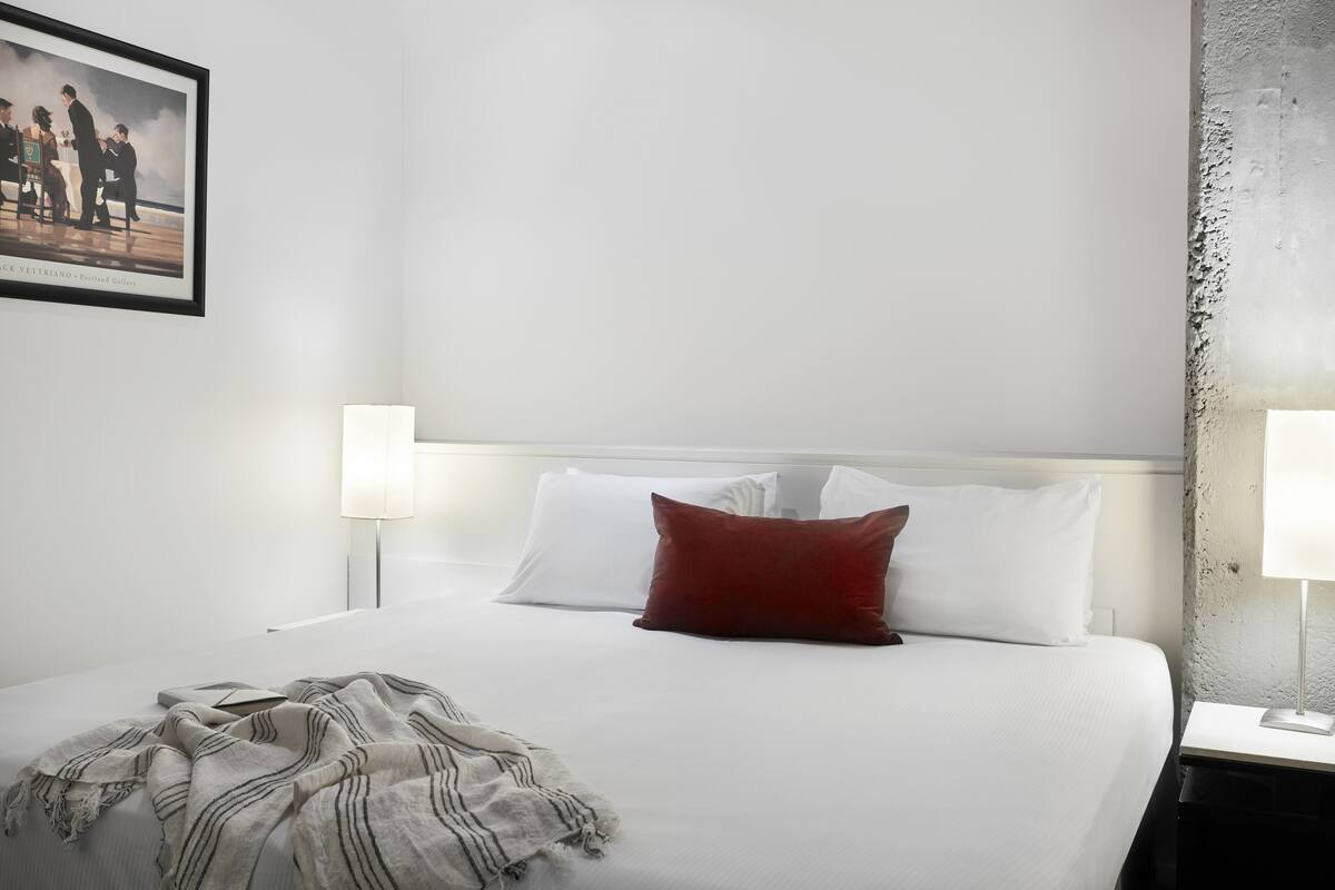 Ein ordentlich bezogenes Bett mit weißen und roten Kissen, einer Streifenwolldecke und einem gerahmten Bild an der Wand.