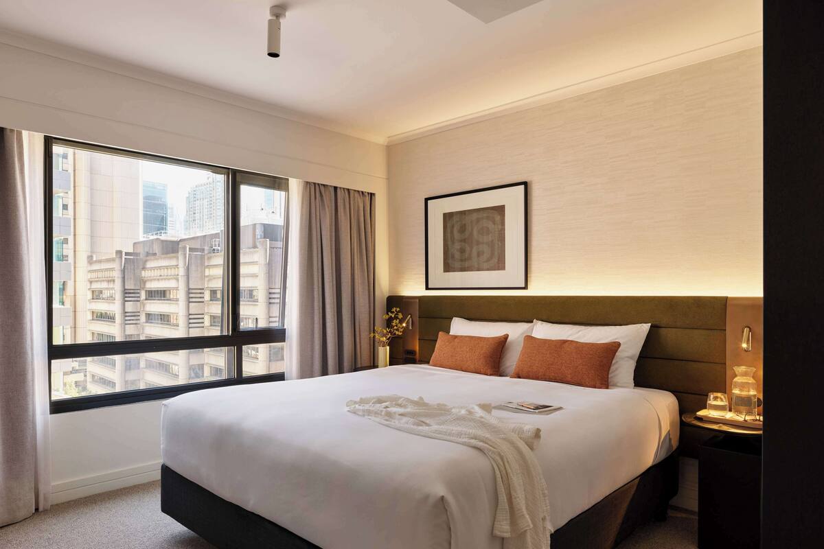Habitación de hotel con una cama grande, una mesita de noche, un cuadro en la pared y una ventana con vista a la ciudad.