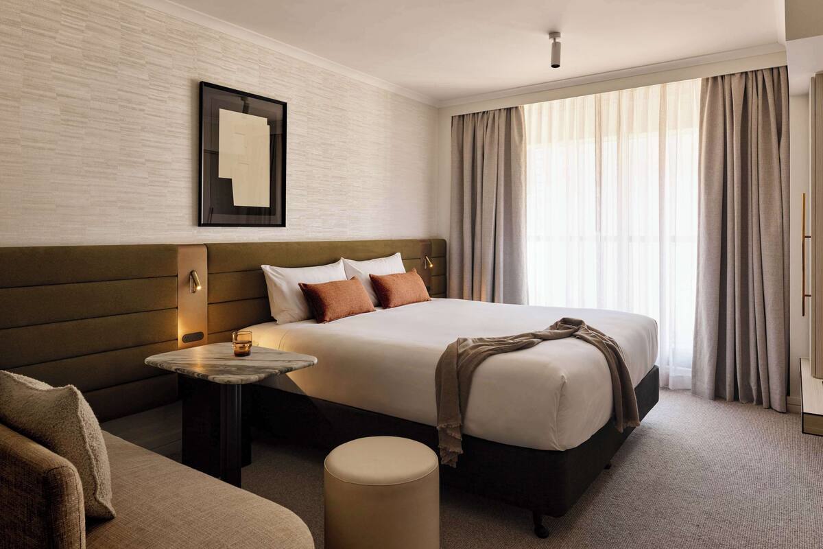 Una habitación de hotel con una cama grande, una mesita de noche con lámpara, un reposapiés redondo pequeño y un cuadro en la pared.