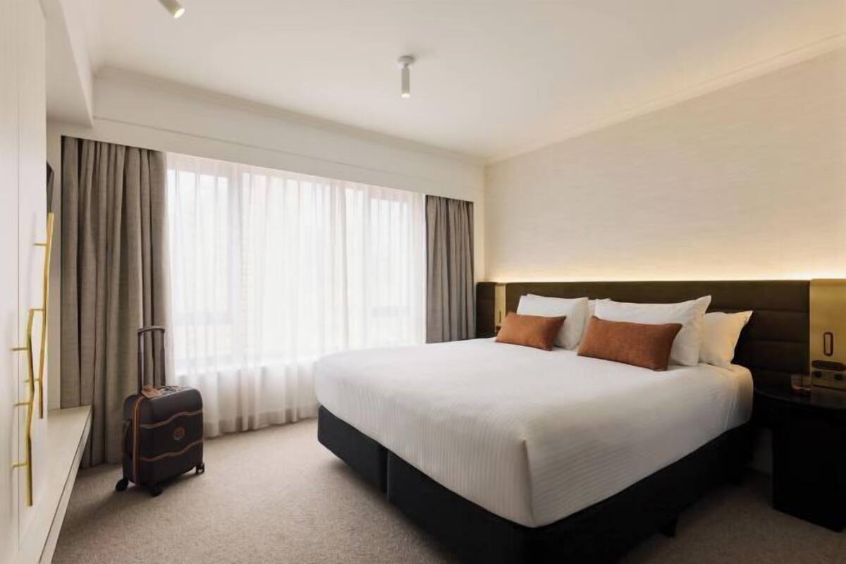 Una habitación de hotel con una cama grande, una mesita de noche, una lámpara, una maleta y una ventana con cortinas.