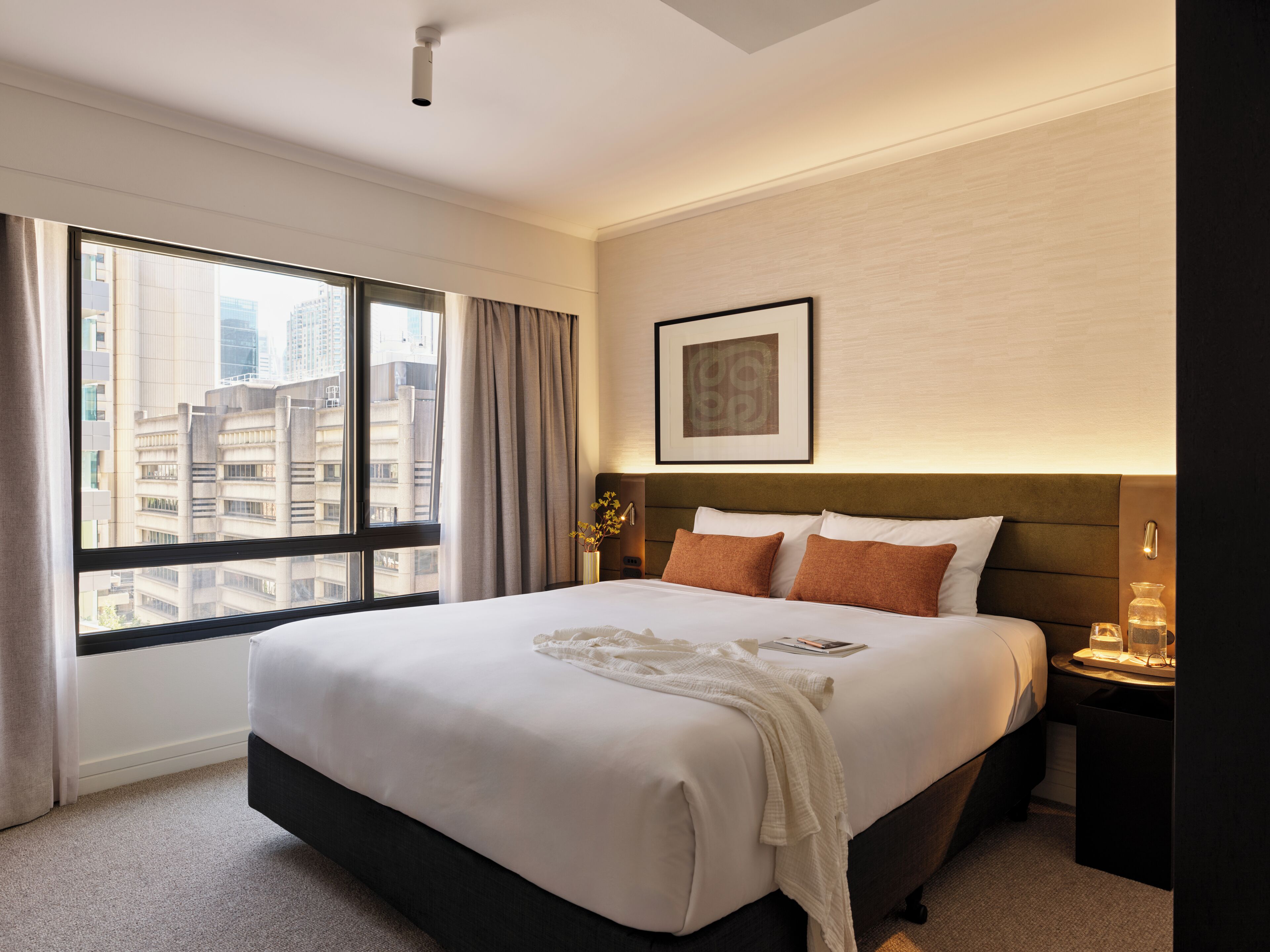 A hotel room with a large bed, a nightstand, a framed picture on the wall, and a view of the city through a large window.