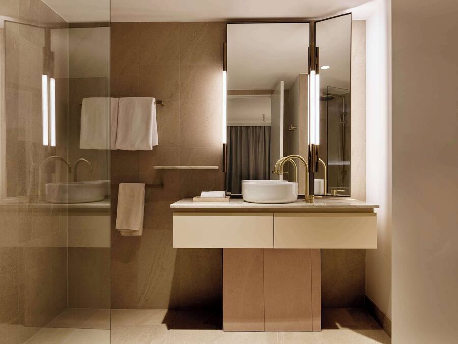 Un baño moderno con ducha envuelta en vidrio, un espejo grande y un lavamanos blanco con detalles dorados.