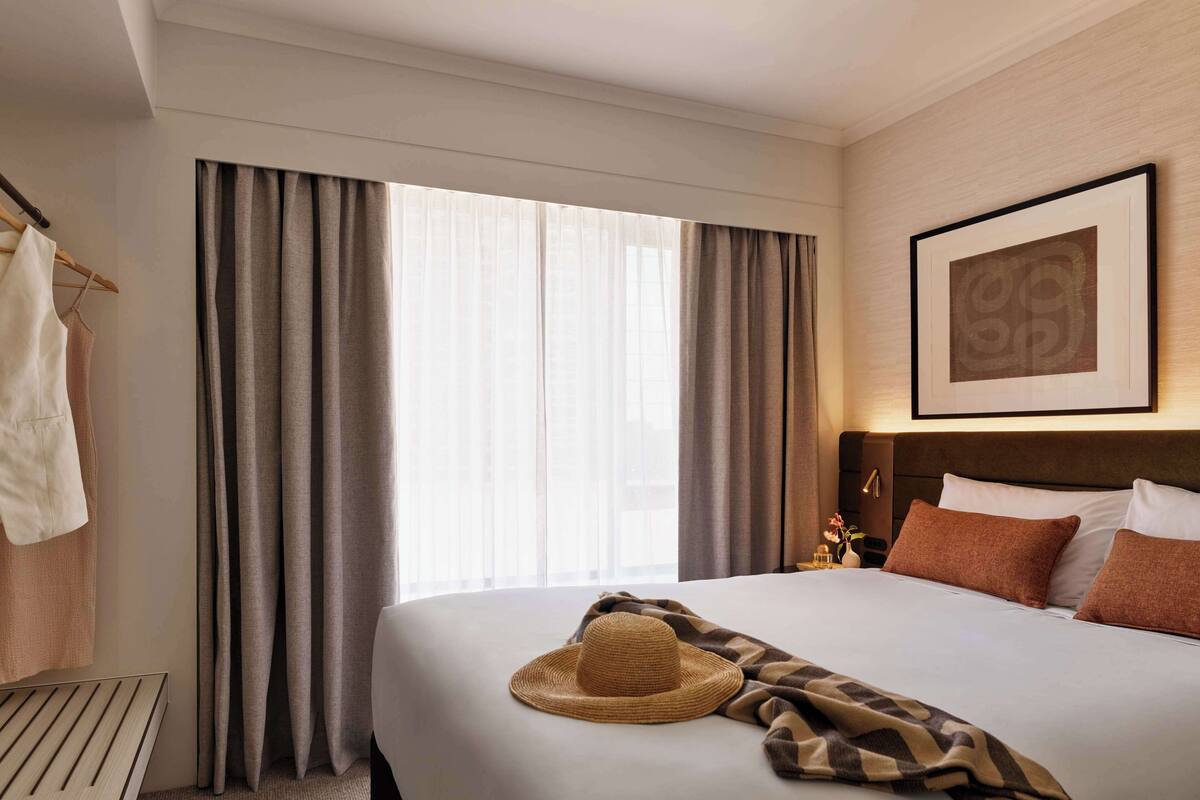 Habitación de hotel con una cama grande, una mesita de noche de madera, un cuadro en la pared y una ventana con cortinas.