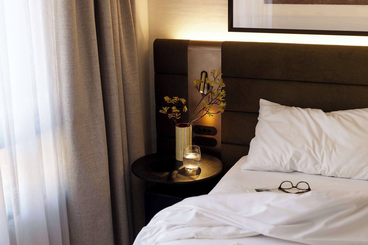 Una cama bien hecha con sábanas blancas, una mesita de noche con un florero y un cuadro en la pared.