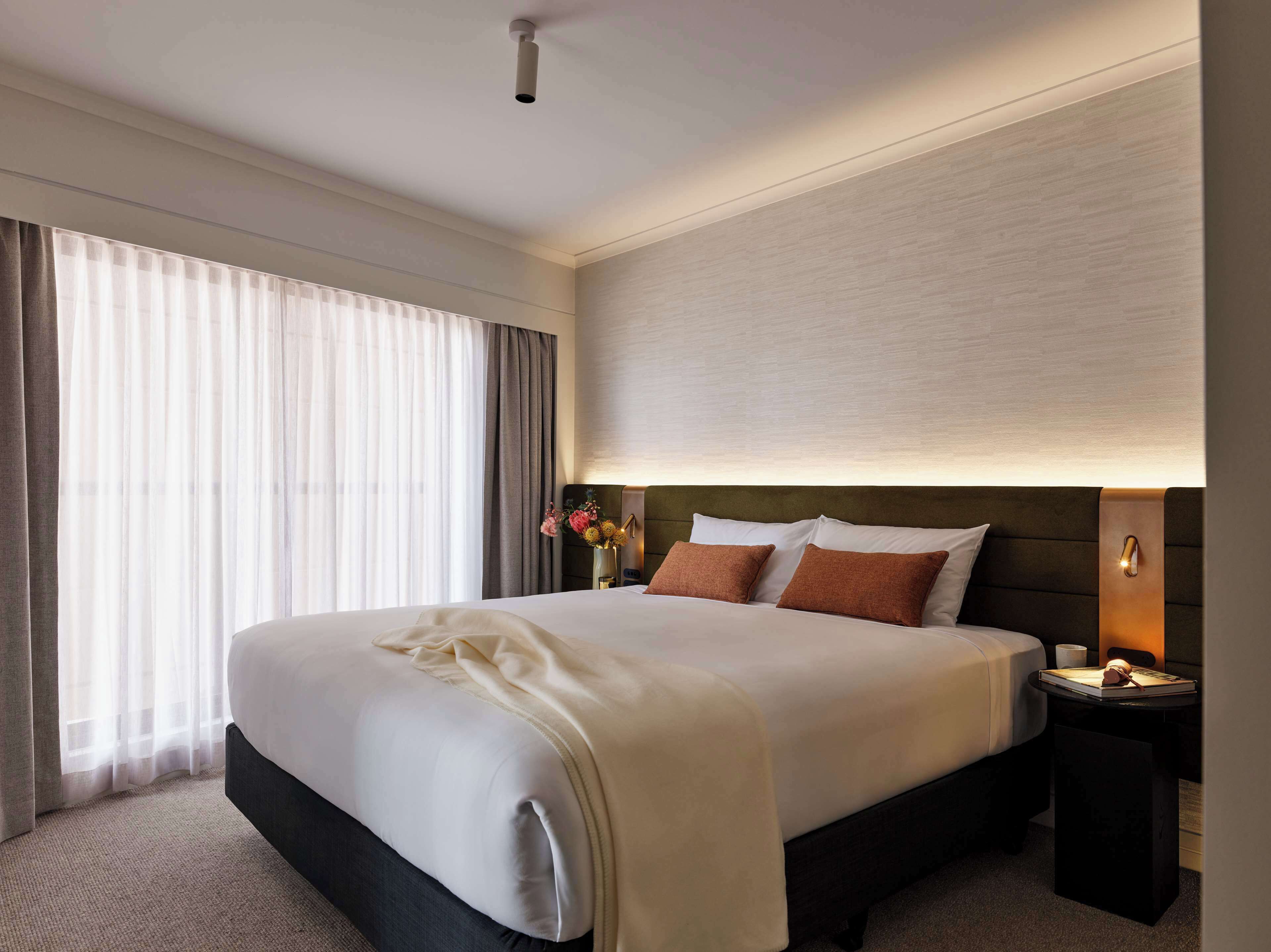 Une chambre d’hôtel avec un grand lit, une table de chevet avec une lampe et une vue sur l’extérieur à travers des rideaux légers.