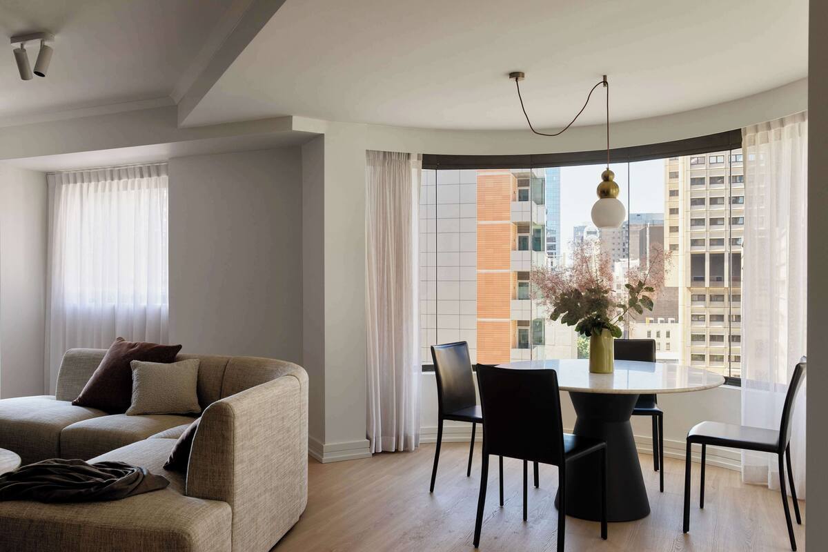 Una sala de estar moderna con sofá, mesa de comedor, sillas y vistas a edificios altos a través de amplios ventanales.